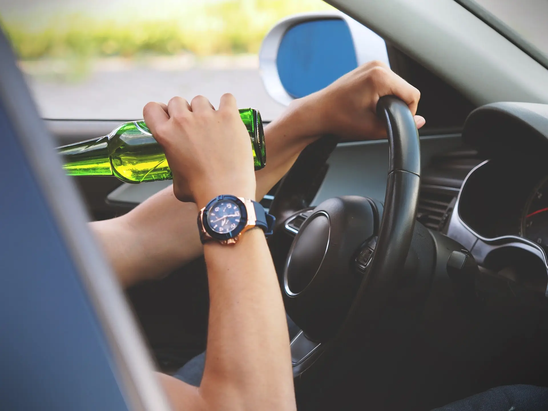 person's left hand on steering wheel and right hand holding a beet bottle to their out-of-frame face