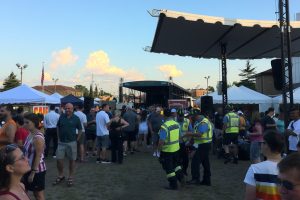 A summer festival during the day, with a lively crowd, food stands, and a music stage.